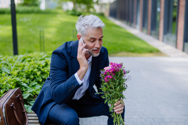 A portrait of happy mature businessman with flower bouquet sitting on bench and waiting for girlfriend.