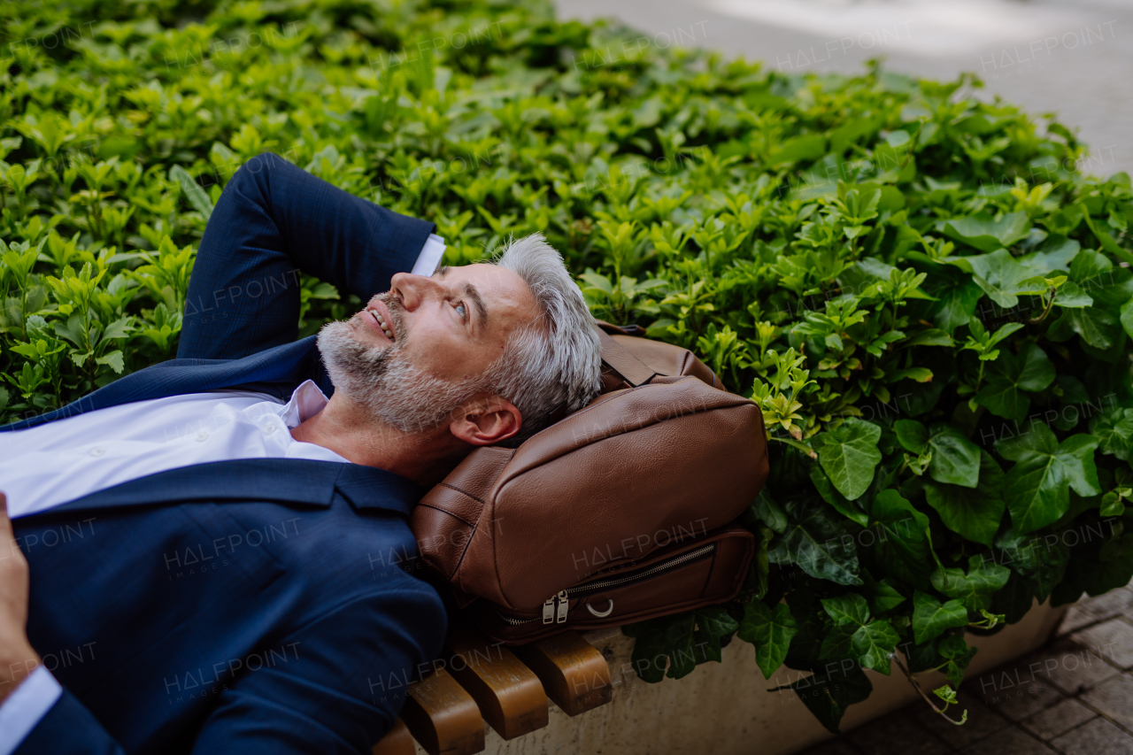 A mature businessman relaxing on bench in city park during break at work, work-life balance concept.