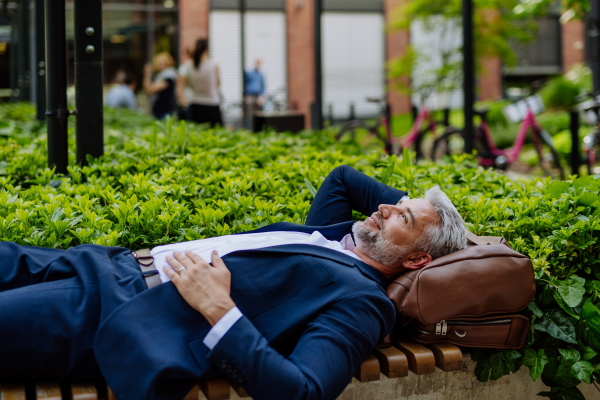 A mature businessman relaxing on bench in city park during break at work, work-life balance concept.