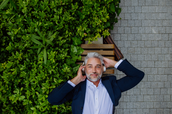 A top view of mature businessman enjoying music in headphones relaxing on bench in city park during break at work.