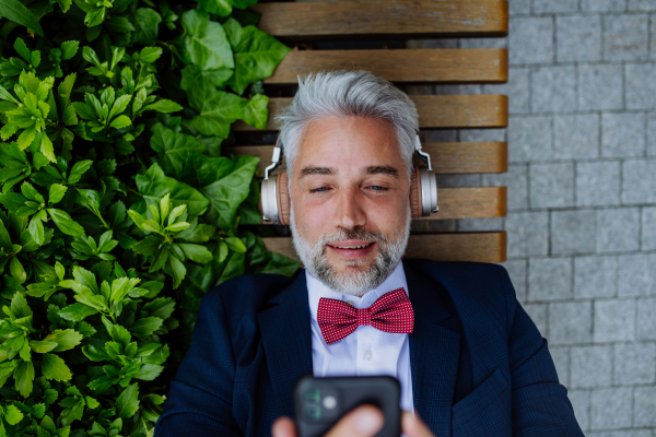 A mature businessman enjoying music in headphones relaxing on bench in city park during break at work.