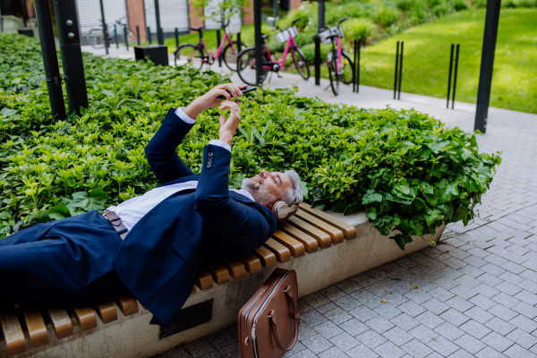 A mature businessman enjoying music in headphones relaxing on bench in city park during break at work.