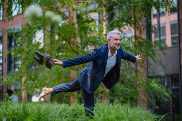 A fun portrait of happy energetic mature businessman carrying shoes and walking barefoot and making pose in park, feeling free, work life balance concept.