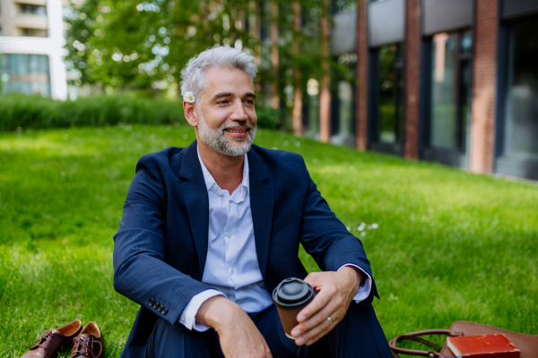 A mature businessman resting and sitting barefoot in park with cup of coffee, feeling free, escaping from work, work life balance concept.