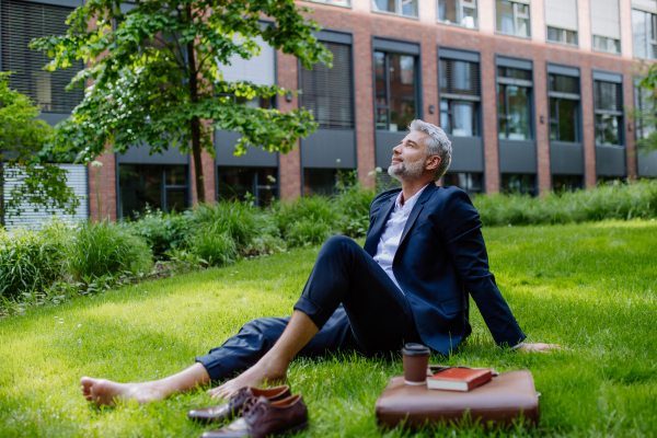 A mature businessman resting and sitting barefoot in park, feeling free, escaping from work, work life balance concept.