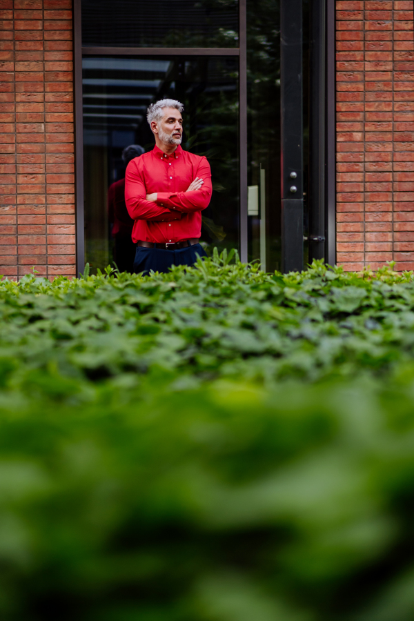 Mature businessman standing in red shirt in office door, next green bushes.