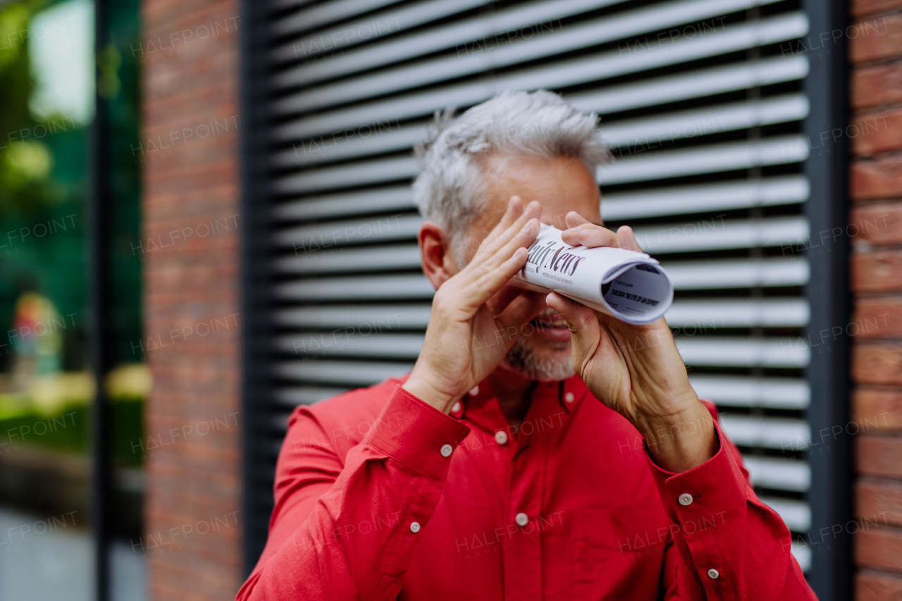 Mature businessman in red shirt looking trough rolling newspaper, standing in the street.