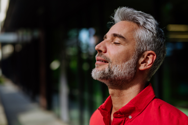 A cheerful mature businessman keeping eyes closed and taking a deep breath while standing outside during a break from work.