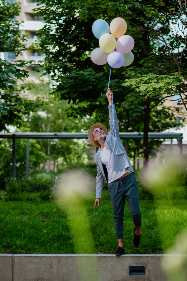 A fun portrait of happy energetic mature businessman holding balloons and walking on wall , feeling free, work life balance concept.