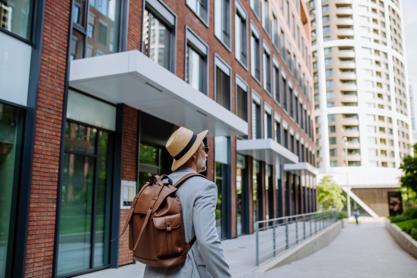 A rear view of man wearing straw hat and backpack walking in street, businessman in casual clothes in summer on the way to work.