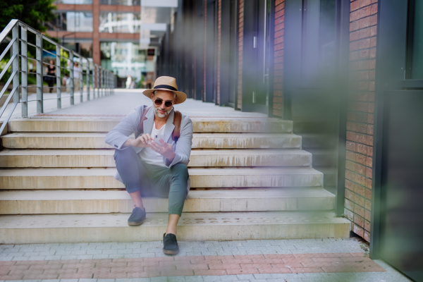 A confident man wearing straw hat and backpack sitting on stairs and using phone, businessman in casual clothes in summer on the way to work.