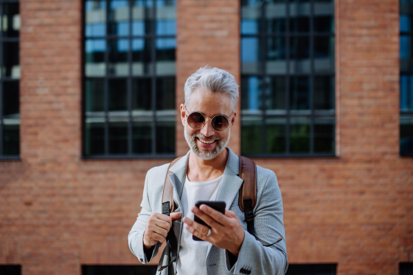 A successful happy businessman waiting in city street, using mobile phone, commuting concept.