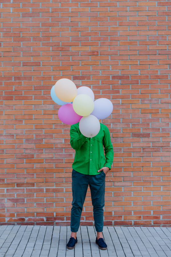 A fun portrait of happy energetic mature man holding balloons in street and hiding behind them, feeling free.