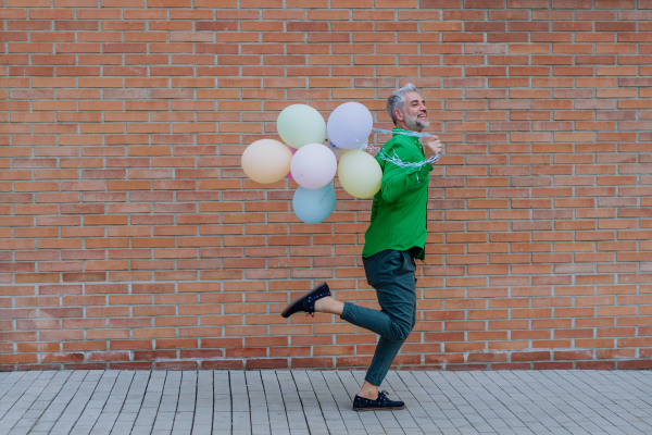 A fun portrait of happy energetic mature man holding balloons and running in street , feeling free.