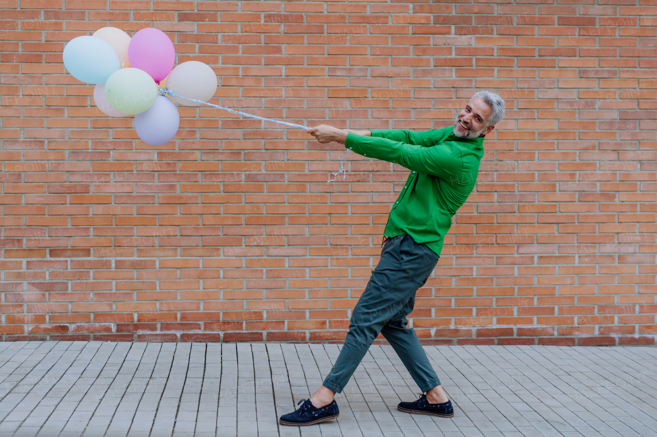 A fun portrait of happy energetic mature businessman holding balloons in street, feeling free, work life balance concept.