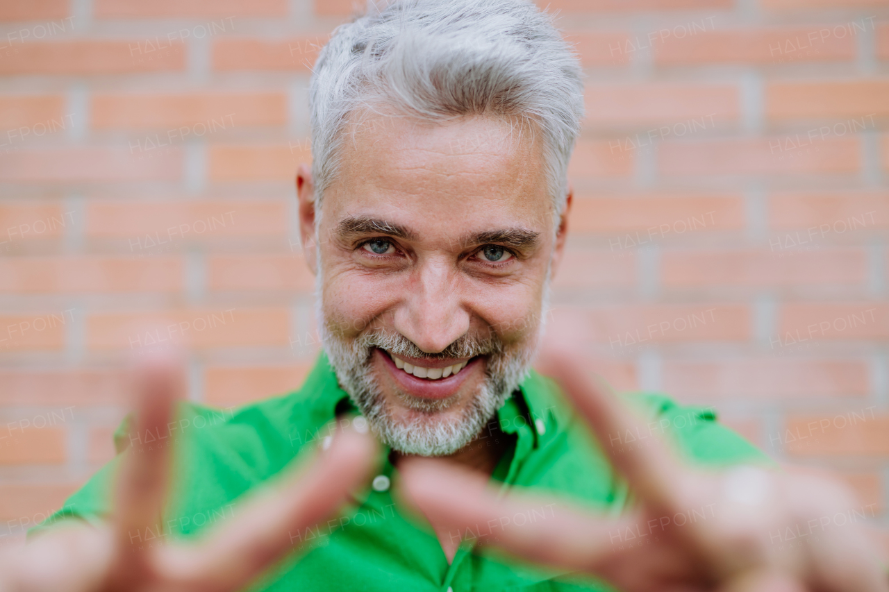 A mature man with happy expression, showing peace sign with fingers and looking at camera, on street background.