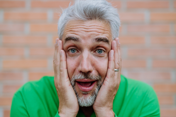 A mature man with shocked, amazed expression touching his face on street background.