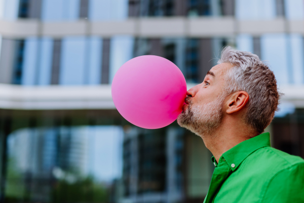 Fun portrait of happy energetic mature man inflating pink balloon in street, standing in front of a skyscraper, copy space.