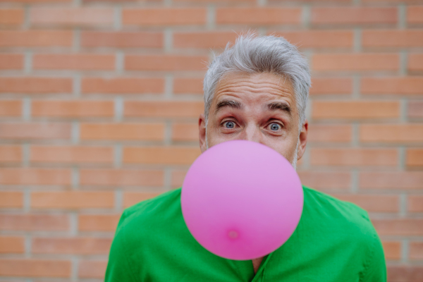 Fun portrait of happy energetic mature man inflating pink balloon in street, standing in front of the brick wall, copy space.