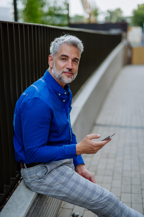 A successful happy businessman waiting in city street, using mobile phone, commuting concept.