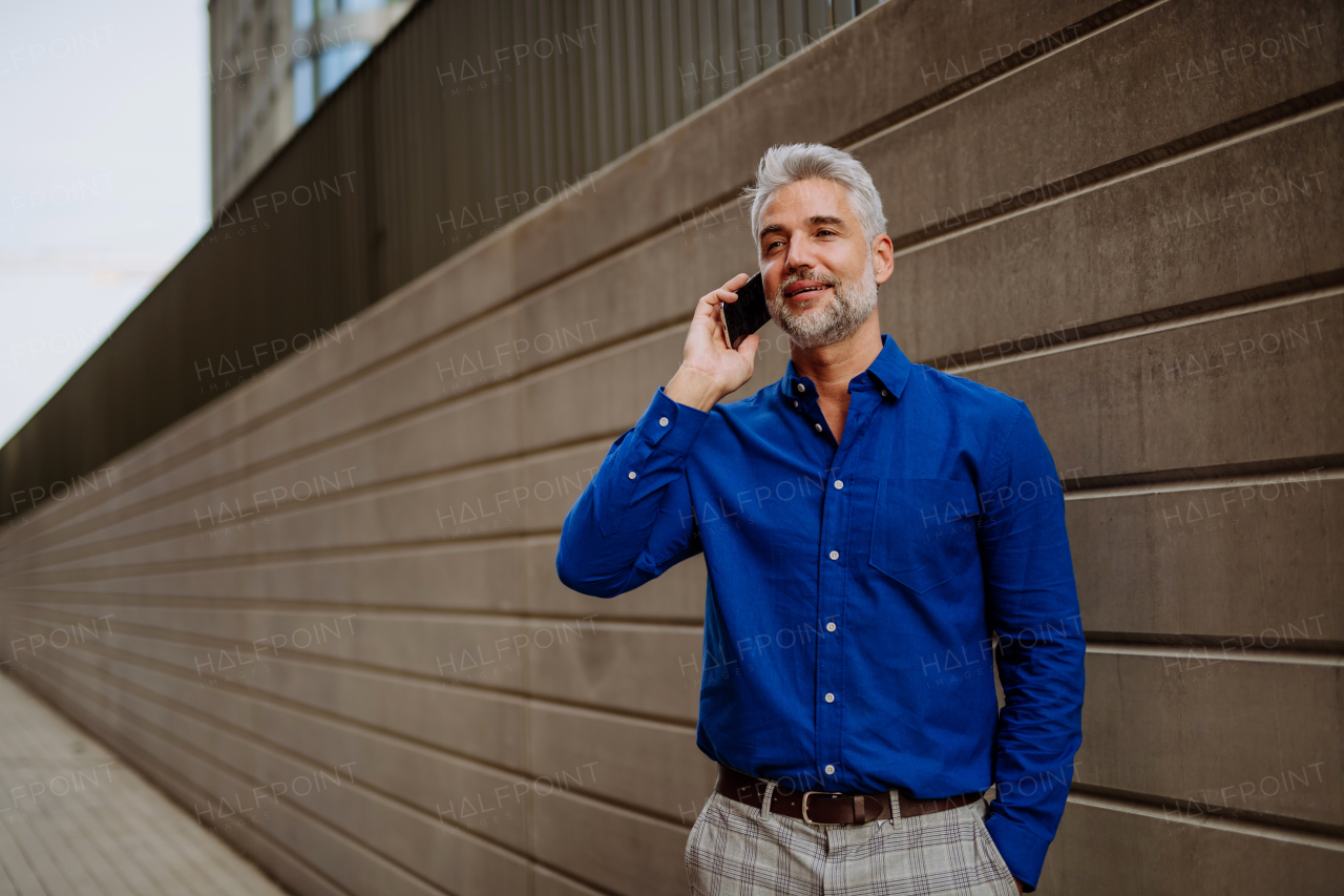 A successful happy businessman waiting in city street, using mobile phone, commuting concept.