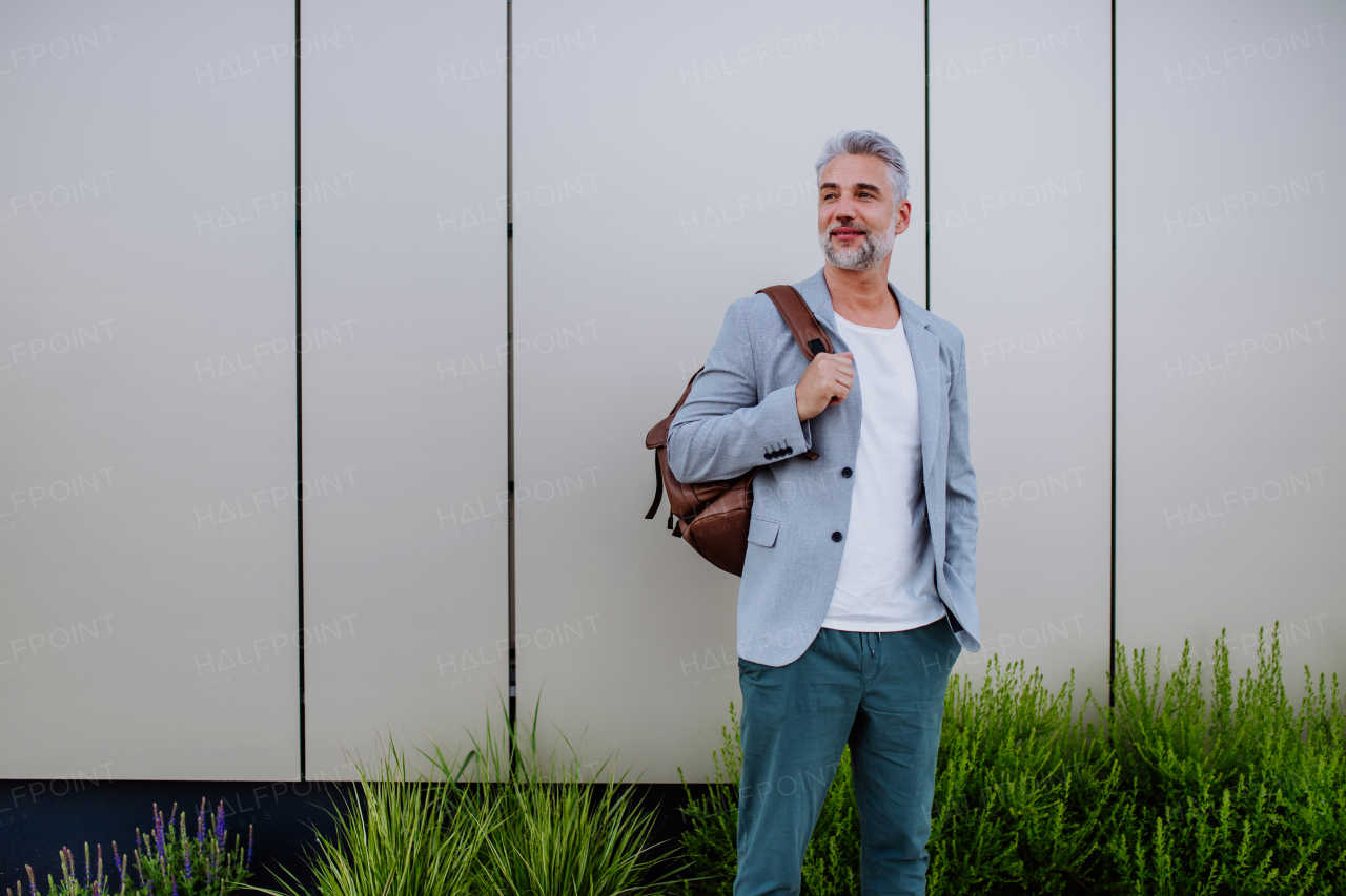 A mature relaxed businessman with backpack leaning on street wall, work life balance concept.