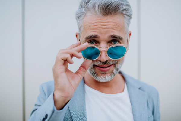 A portrait of successful happy businessman wearing causal clothes and sunglasses in summer in street looking at camera.