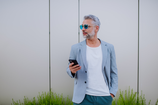 A successful happy businessman waiting in city street, using mobile phone, commuting concept.