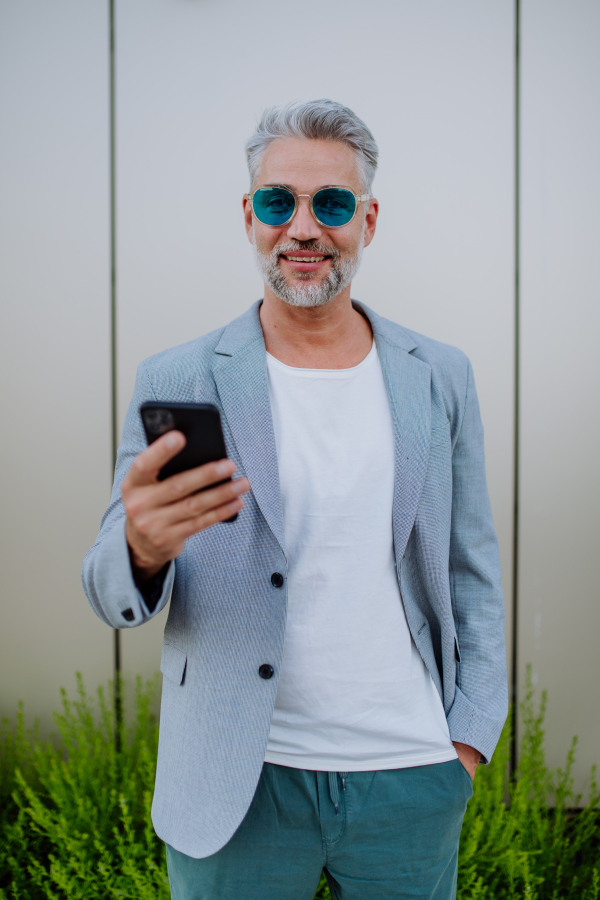 A successful happy businessman waiting in city street, using mobile phone, commuting concept.