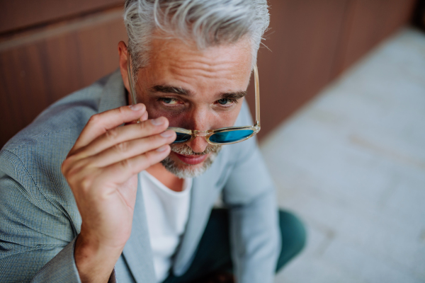 A portrait of successful happy businessman wearing causal clothes and sunglasses in summer in street looking at camera.