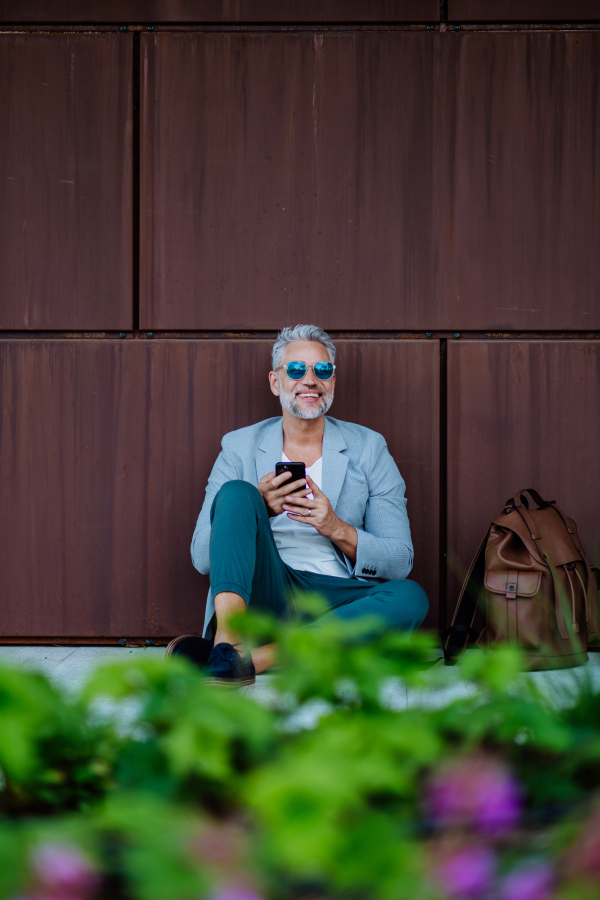 Mature businessman sitting with smartphone in his hand and relaxing in flower city park during a break at work.