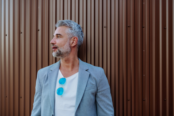 A portrait of uccessful happy businessman wearing causal clothes and sunglasses in summer in street looking away.