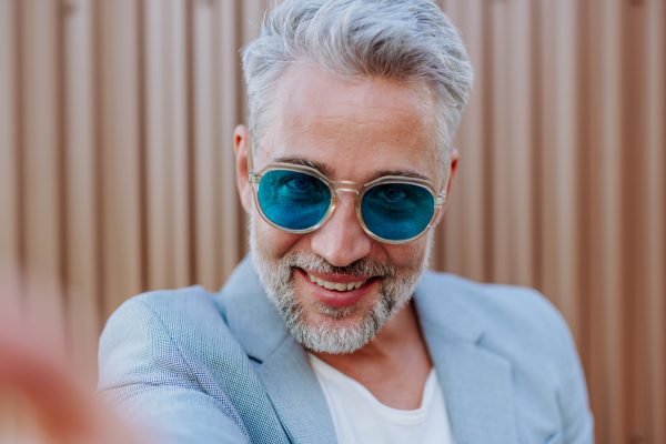 A portrait of successful happy businessman wearing causal clothes and sunglasses in summer in street looking at camera.