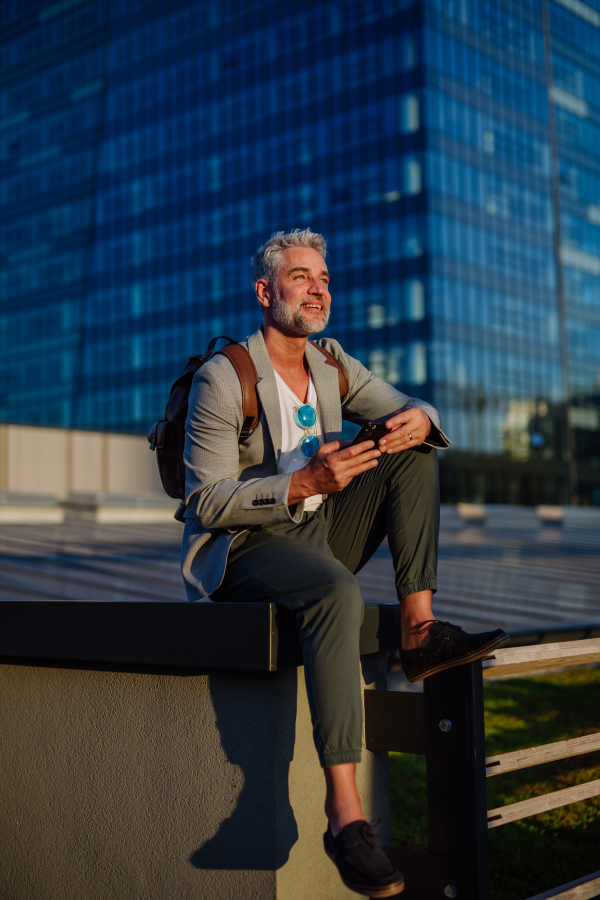 A confident businessman with backpack sitting on stairs and using phone, feeling free, escaping from work, work life balance concept.