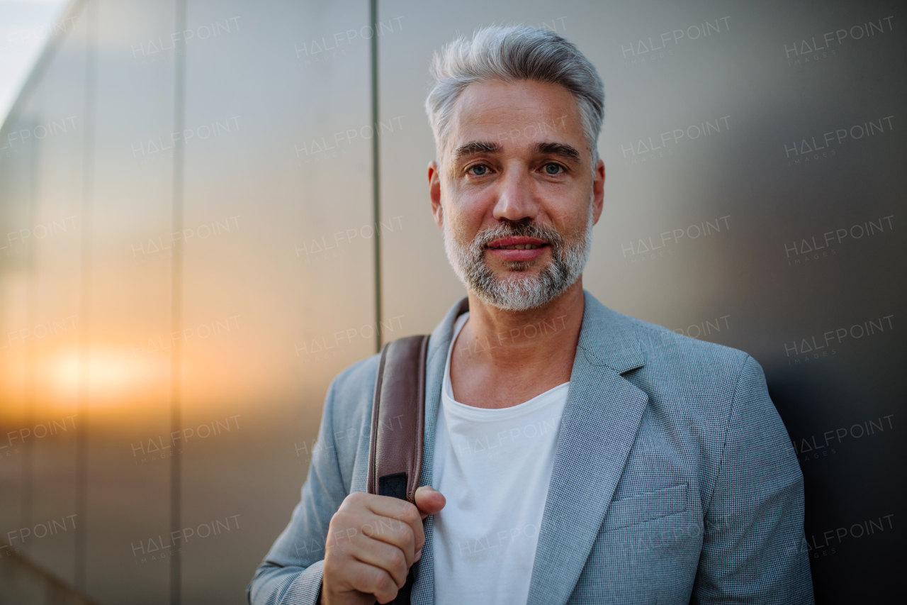 A mature relaxed businessman leaning on street wall and looking at camera, work life balance concept.