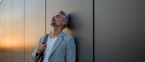 A mature relaxed businessman with closed eyes leaning on street wall and daydreaming, wotk life balance concept.