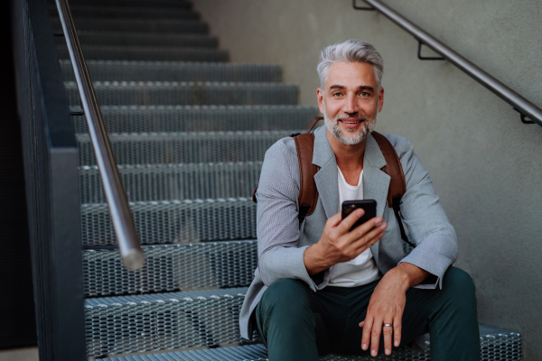 A successful happy businessman waiting in city street, using mobile phone, commuting concept.