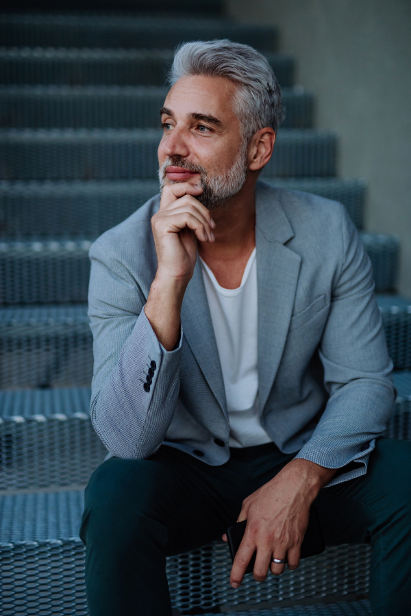 A successful happy businessman waiting in city street, sitting on stairs, commuting concept.