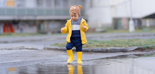 Happy little boy jumping in puddle after rain in cold autumn day.