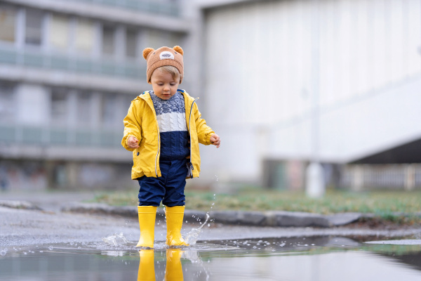 Happy little boy in yellow raincoat jumping in puddle after rain in cold autumn day.