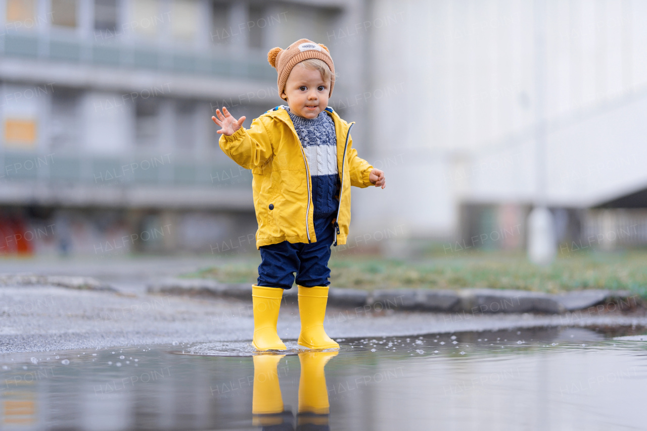 Happy little boy in yellow raincoat jumping in puddle after rain in cold autumn day.