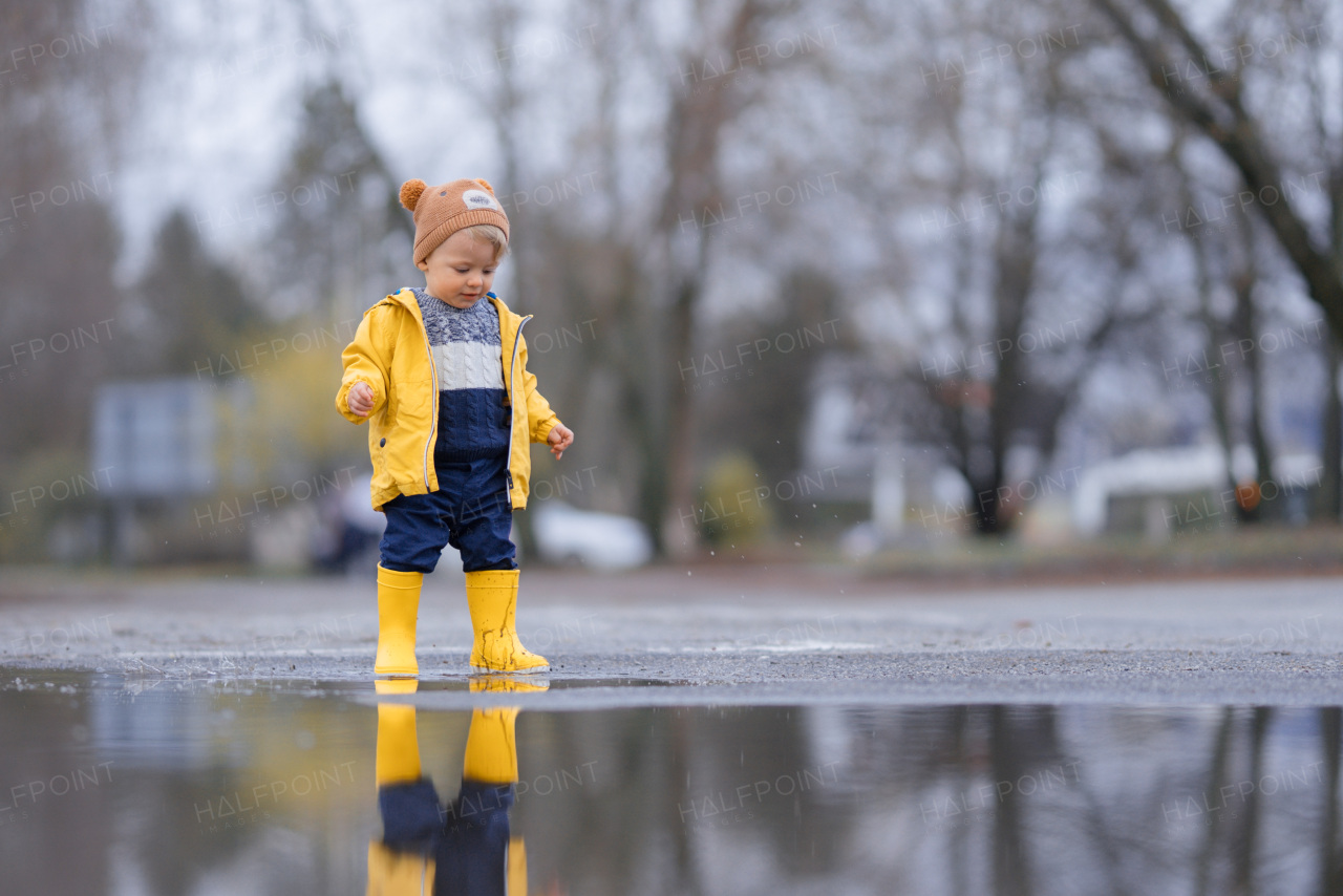 Happy little boy in yellow raincoat jumping in puddle after rain in cold autumn day.