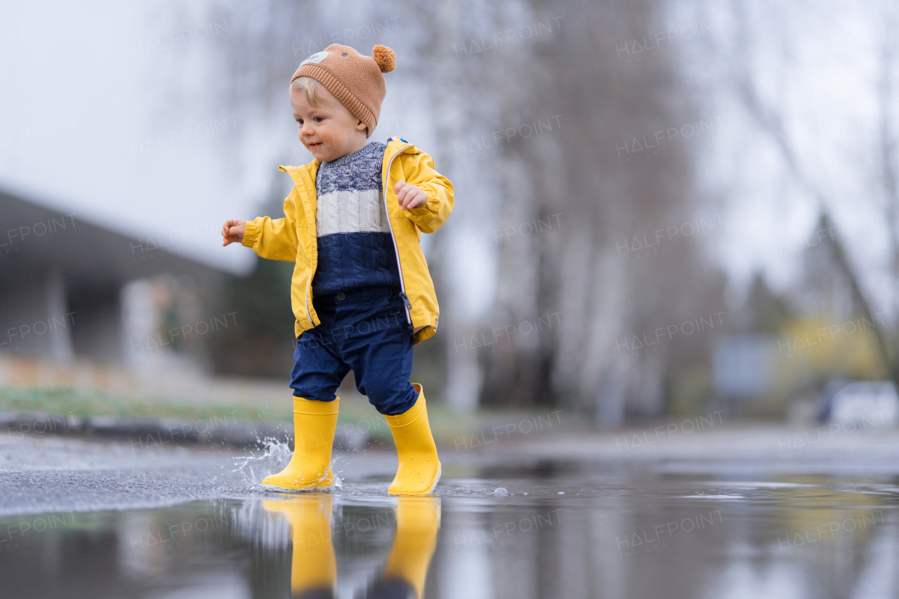 Happy little boy in yellow raincoat jumping in puddle after rain in cold autumn day.