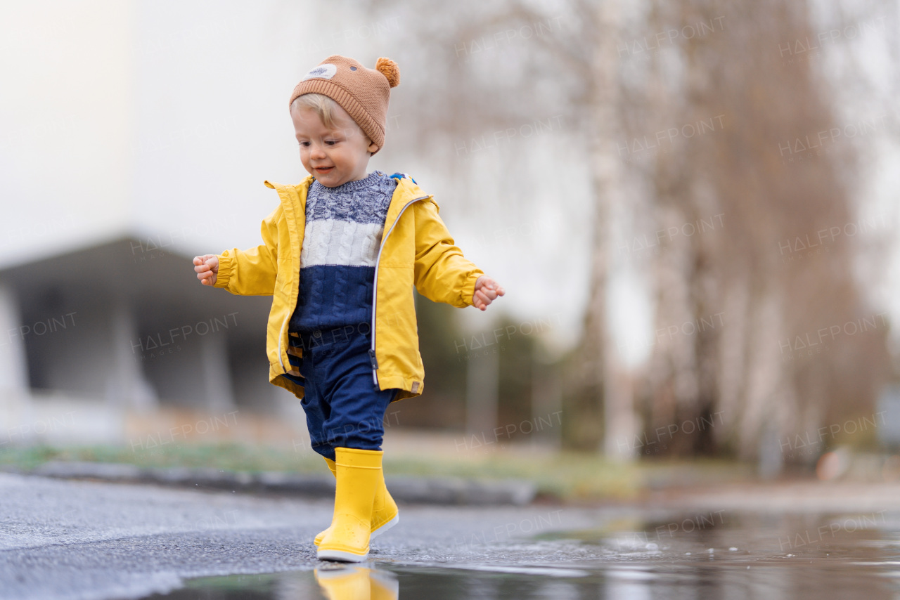 Happy little boy in yellow raincoat jumping in puddle after rain in cold autumn day.