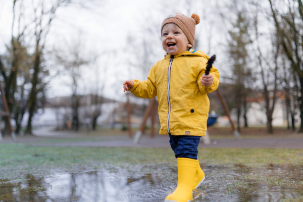 Happy little boy in yellow raincoat jumping in puddle and playing with conifer cone after rain.