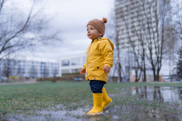 Happy little boy in yellow raincoat jumping in puddle after rain in cold autumn day.