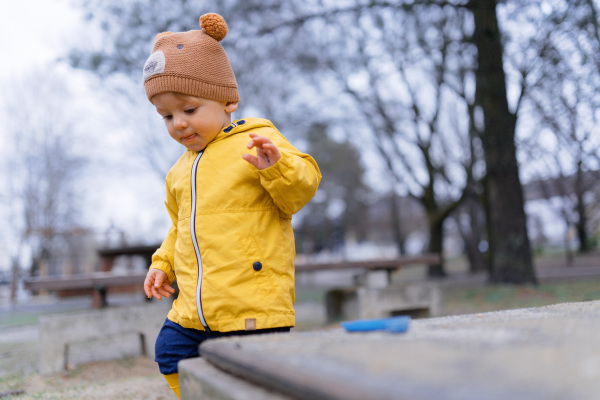 Happy little boy with a teddy bear cap playing on playground at sandpit during autumn day.