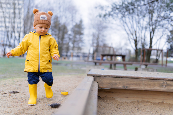 Happy little boy with a teddy bear cap playing on playground at sandpit during autumn day.