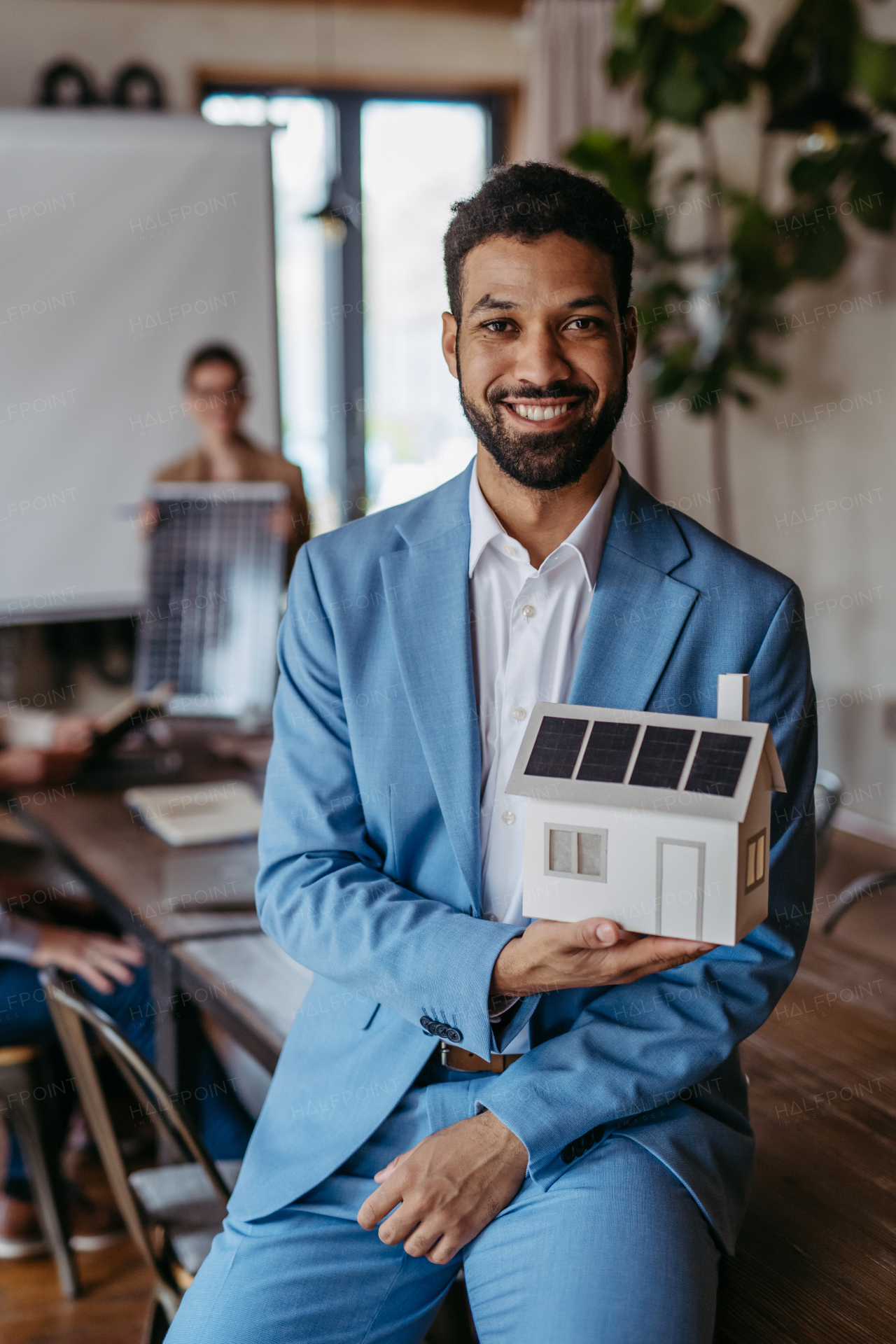 Portrait of confident young businessman holding model of house with solar panels in meeting room. Young colleagues discussing photovoltaic panels, clean, green, energy. Business using renewable energy.