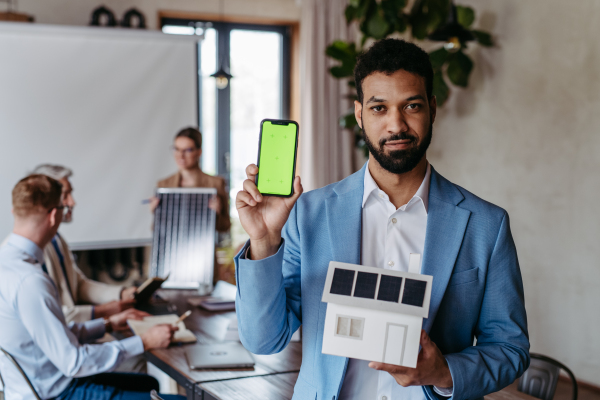 Portrait of confident young businessman holding model of house with solar panels, showing screen of smartphone. Young colleagues discussing photovoltaic panels, clean, green, energy. Mobile phone mockup screen with blank space.